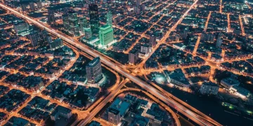 An aerial drone photograph of a city at twilight, showing geometric patterns of streetlights and building illumination in amber and green, symbolizing macroeconomic forces shaping urban life. The image represents the interconnectedness of economic indicators with daily financial well-being.