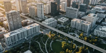 Aerial view of a city at dawn, showing geometric buildings, light rail, and a large green community park, symbolizing the intersection of economic development and social well-being.